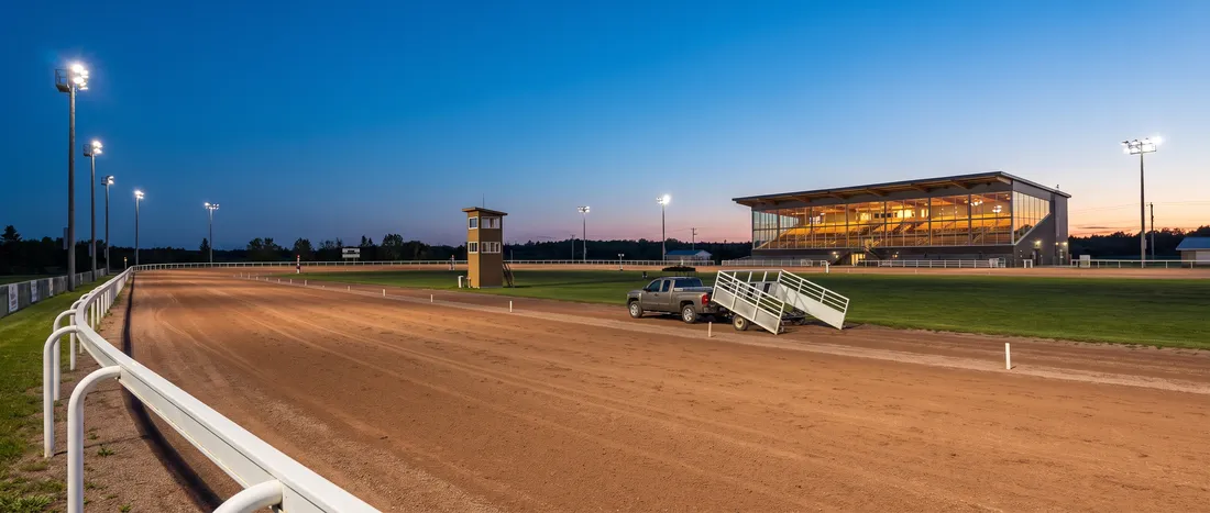 Illustration: an empty harness racing oval at blue hour, a white rail, a lit two-storey grandstand, a slim judges tower in the infield and a starting gate truck on the track.